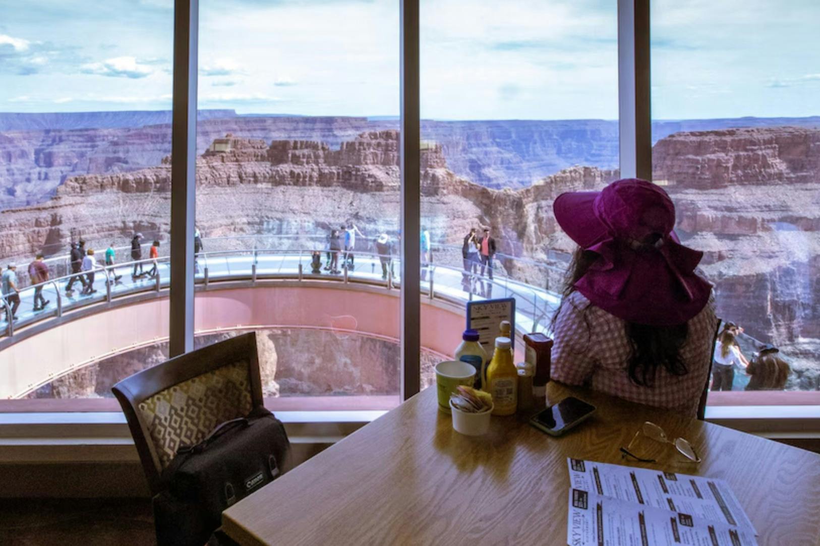 A person seated indoors near a window overlooking the Grand Canyon Skywalk and the scenic canyon view beyond.