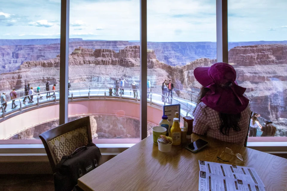 A person seated indoors near a window overlooking the Grand Canyon Skywalk and the scenic canyon view beyond.
