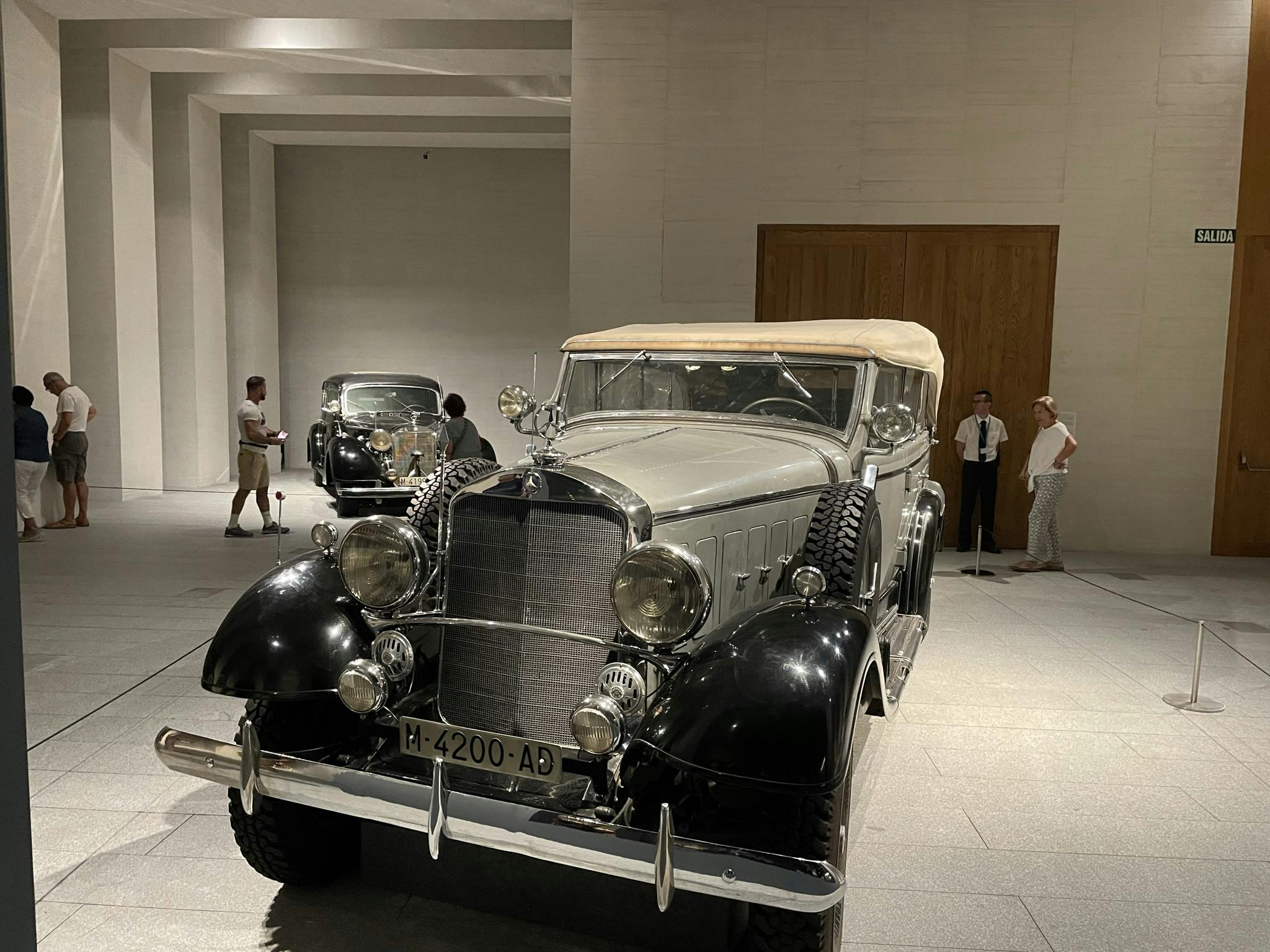 室内陈列着两辆老爷车，人们在旁观看。 前车有米色软顶和黑色挡泥板。