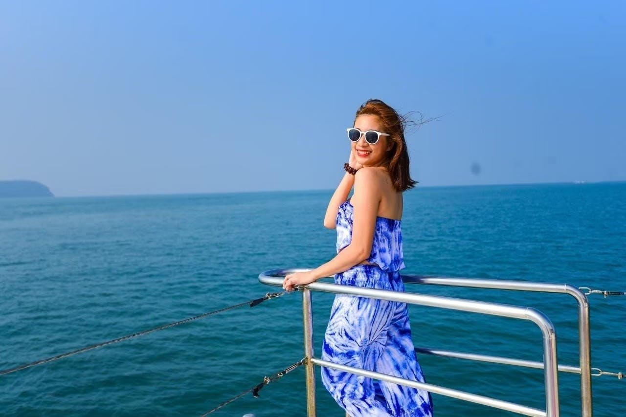 一位身穿蓝色连衣裙、戴着白色太阳镜的女士在船栏杆上微笑，背景是大海和晴朗的蓝天。