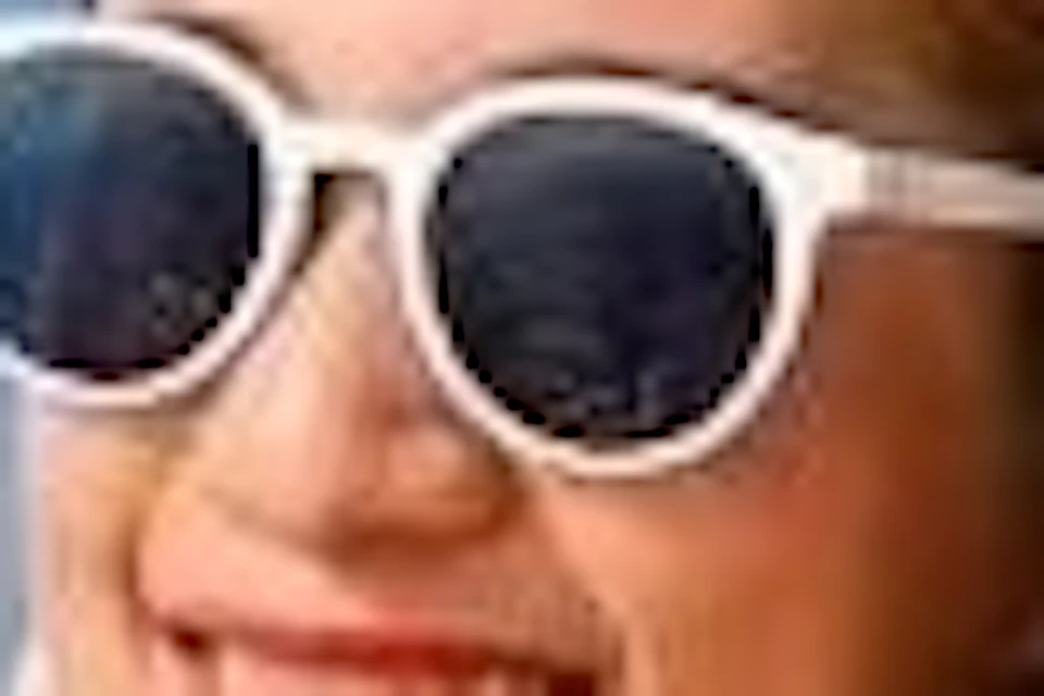 Woman in a blue dress and white sunglasses smiles on a boat's railing, with the sea and a clear blue sky in the background.