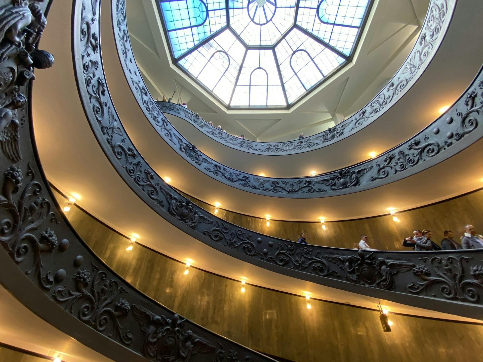 Curved staircases with ornate railings spiral upward inside a building, under a large, geometric skylight. People are visible on the stairs.