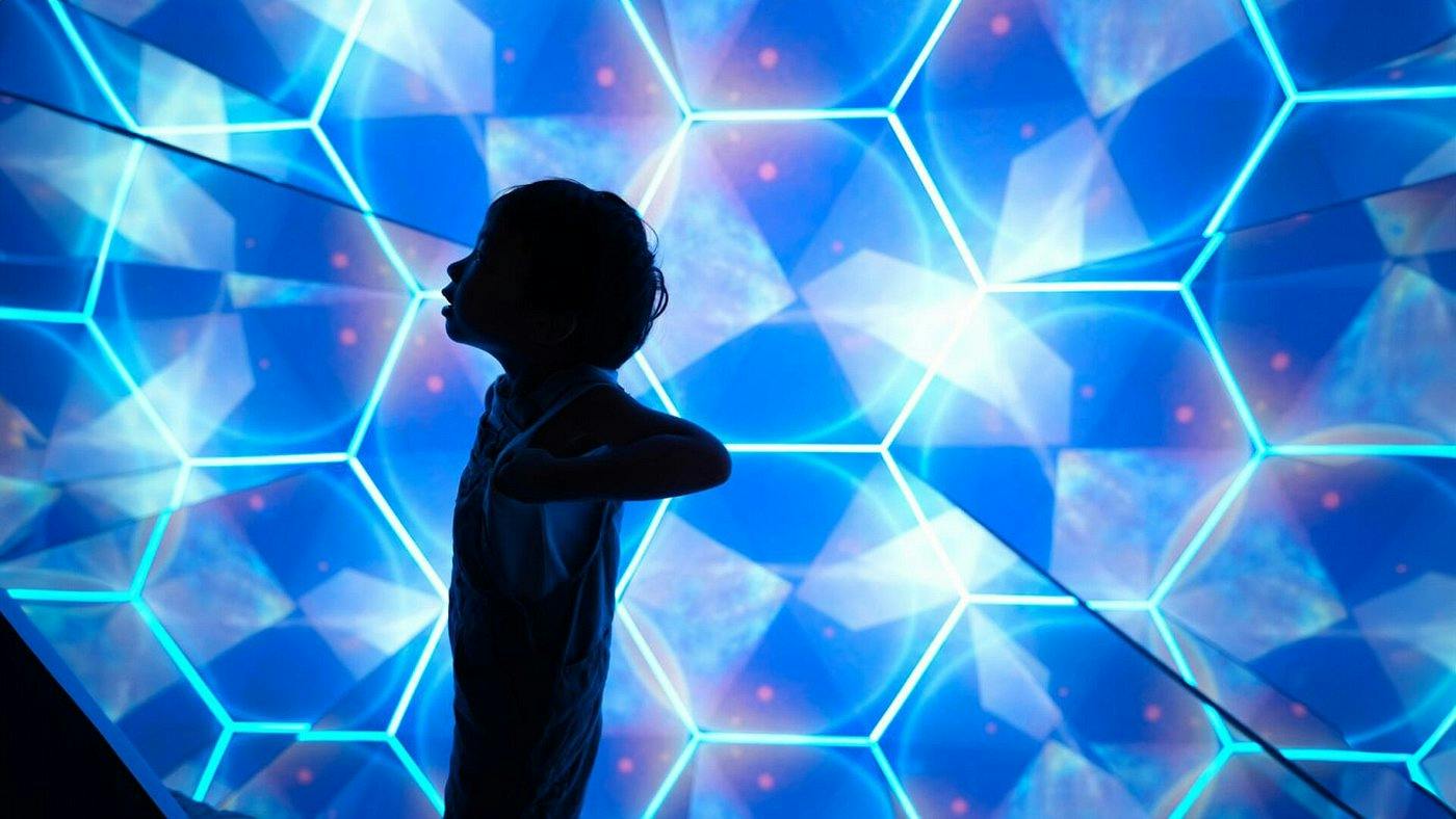 Silhouette of a child in front of a vibrant geometric blue light display with glowing hexagonal patterns.