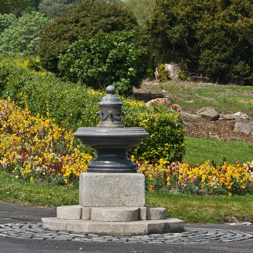 Stenvattenfontän i en park, omgiven av en blomsterbädd med gula och röda blommor och frodiga gröna buskar och träd.