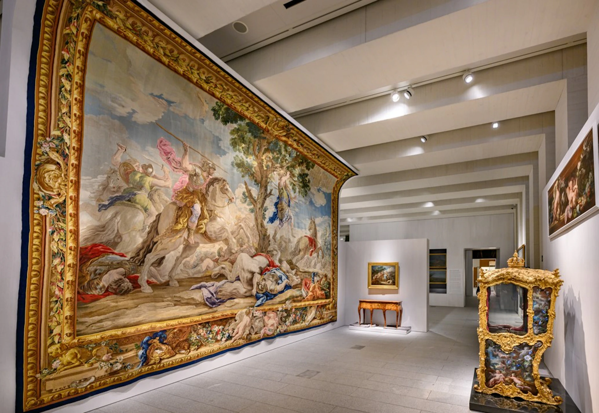 A museum gallery displaying a large, ornate tapestry depicting a battle scene, with a smaller framed painting on the back wall.