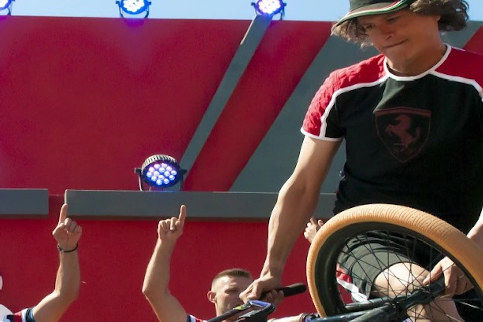 A BMX rider performs a stunt on a stage while four dancers in red pants and white shirts cheer in the background.