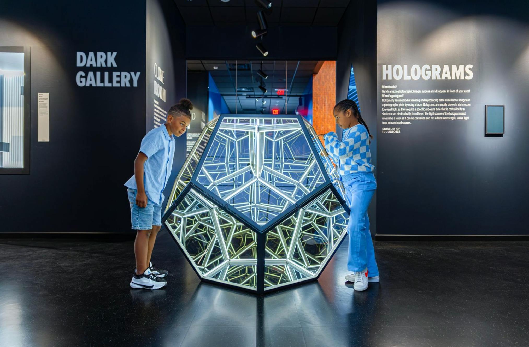 Zwei Kinder betrachten eine leuchtende geometrische Skulptur in einer dunklen Galerie. An den Wänden befinden sich Texte zu den Exponaten, darunter auch Hologramme.