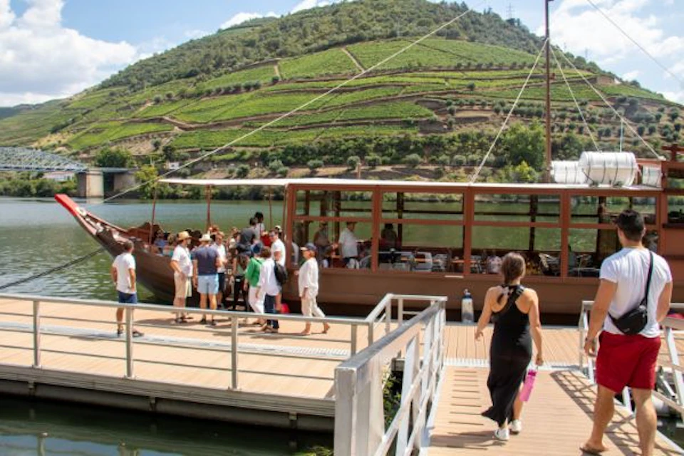 People boarding a wooden boat at a dock with a vineyard-covered hill in the background under a clear blue sky.