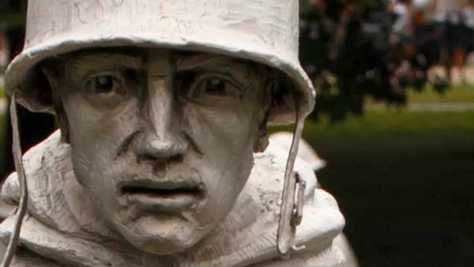 Statues of soldiers in rain cloaks and helmets stand among shrubs and trees, depicting a military scene in a memorial setting.