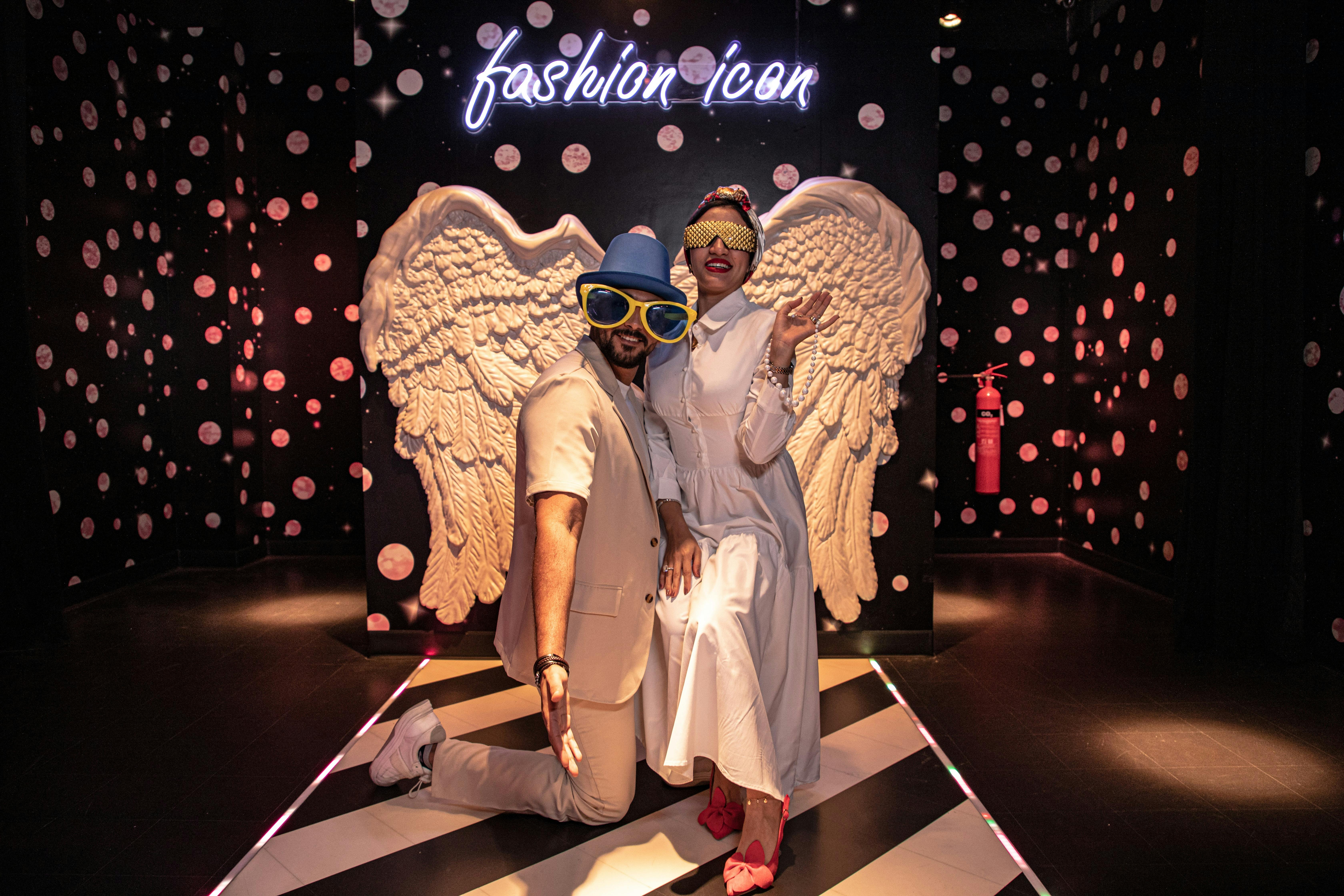 Před křídlovou sochou pózují dva lidé v nadrozměrných brýlích. Neonové a puntíkované pozadí čte „módní ikona“.