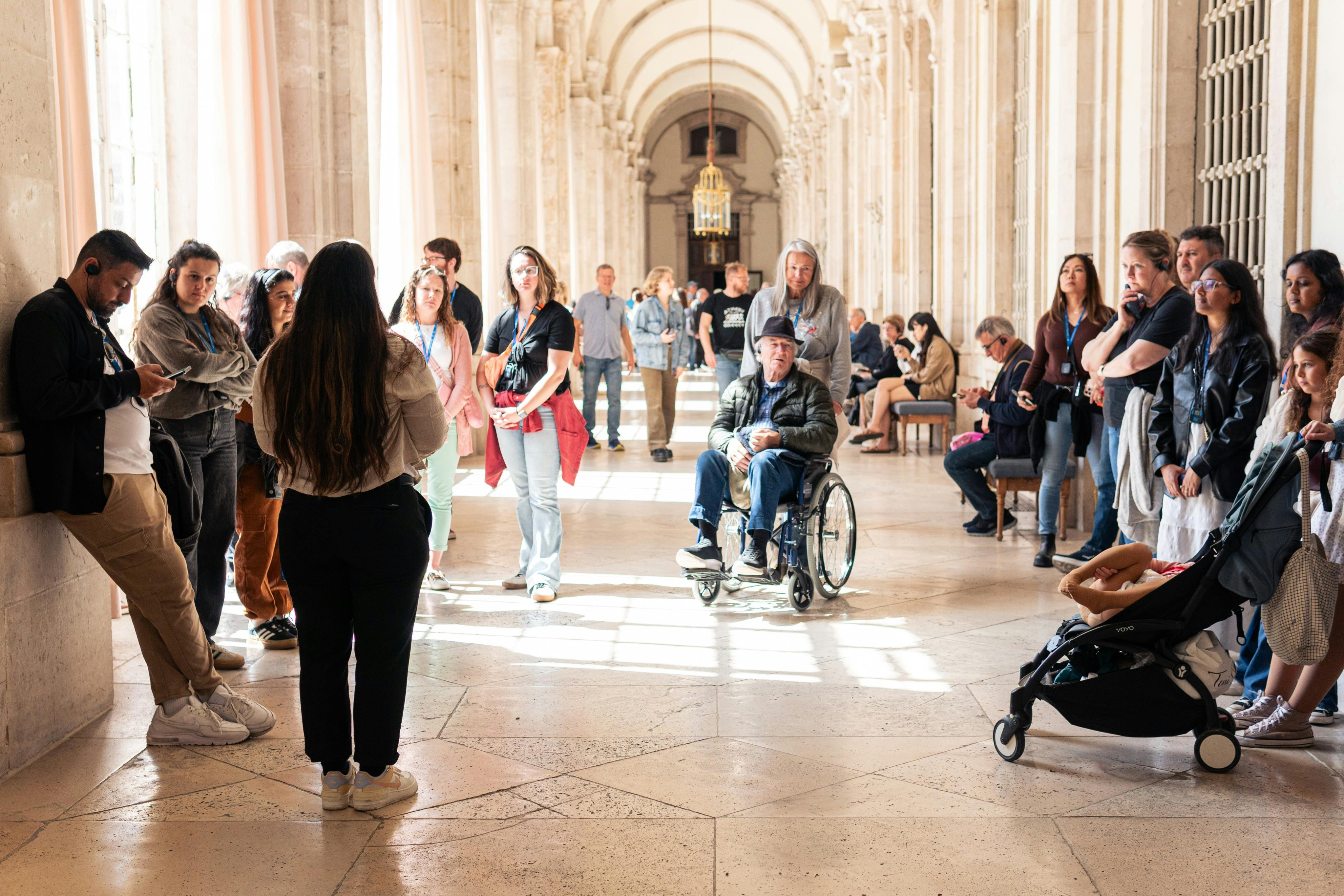 A group of people in a bright, arched hallway, including a man in a wheelchair and a person with a stroller.