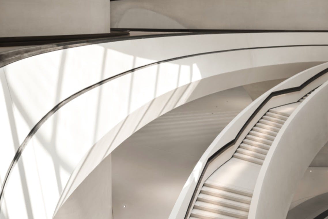 Zakřivený bílý architektonický design se schody a zábradlím, osvětlený stíny vrhajícími přirozené světlo.