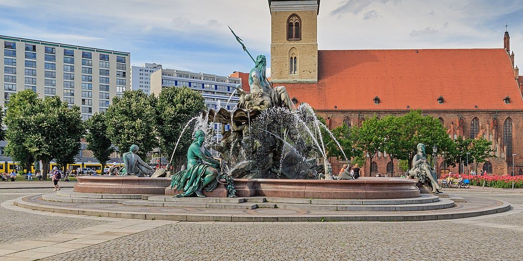 Stone fountain with statues, spouting water jets, set in a plaza with people around. Background features modern and old buildings.
