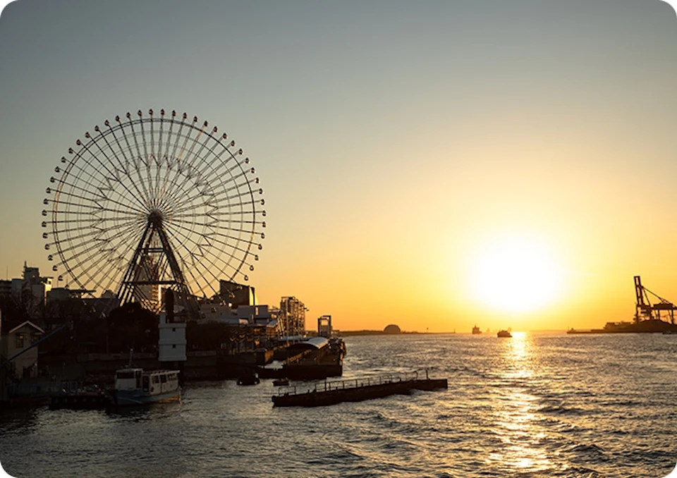 A Ferris wheel near a waterfront at sunset with a clear sky and boats visible on the water.