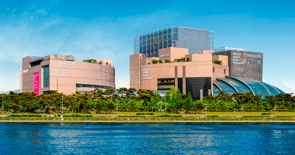Modern buildings with beige facades and greenery on rooftops, located near a river with trees in the foreground.