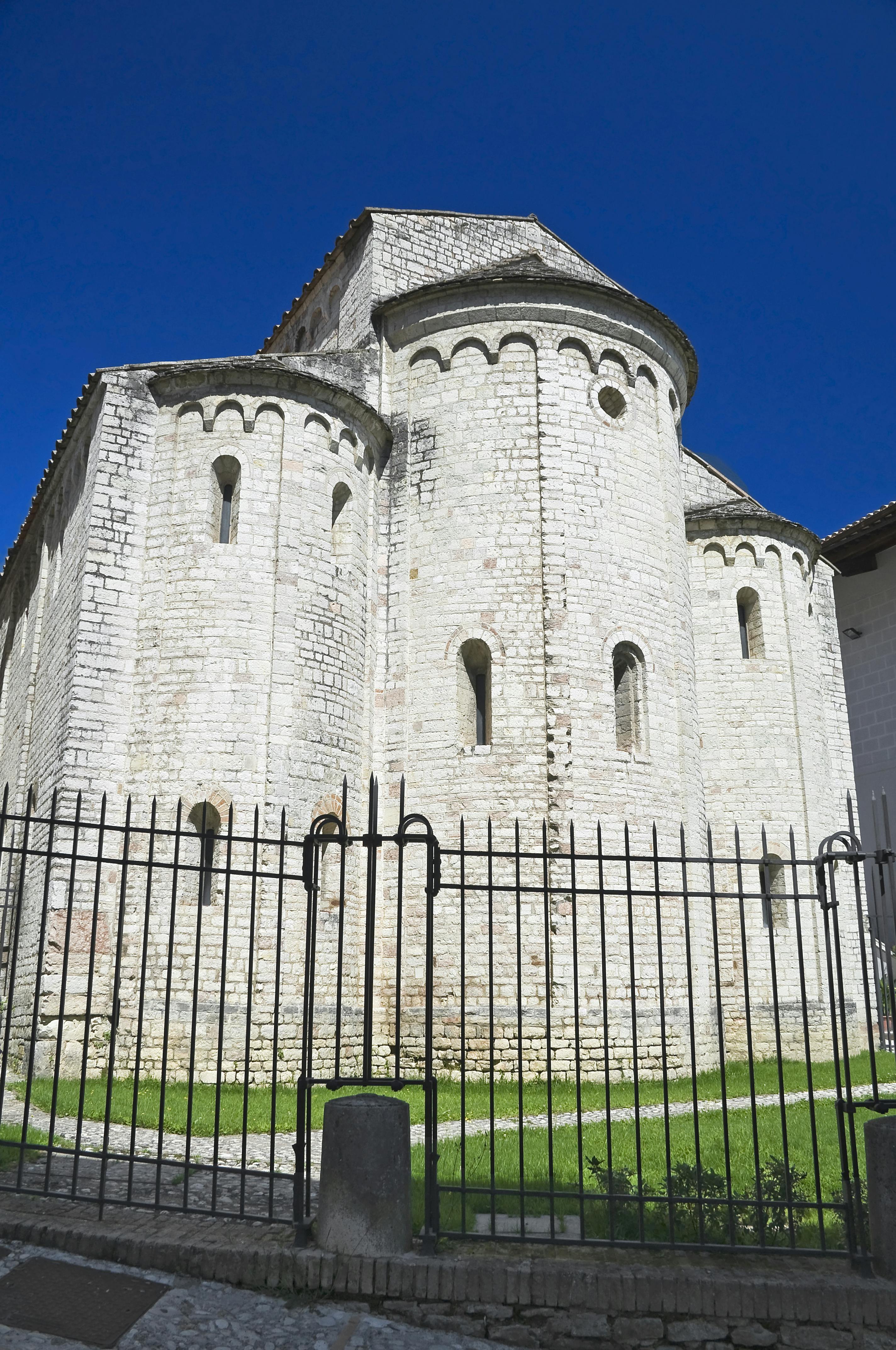 Μια πέτρινη εκκλησία με στρογγυλεμένες αψίδες και στενά παράθυρα, που περιβάλλεται από έναν μαύρο μεταλλικό φράχτη κάτω από έναν καταγάλανο ουρανό.