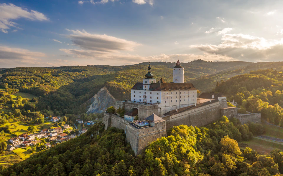Hilltop castle with white walls and multiple towers, surrounded by lush green forests and a village visible in the valley below.