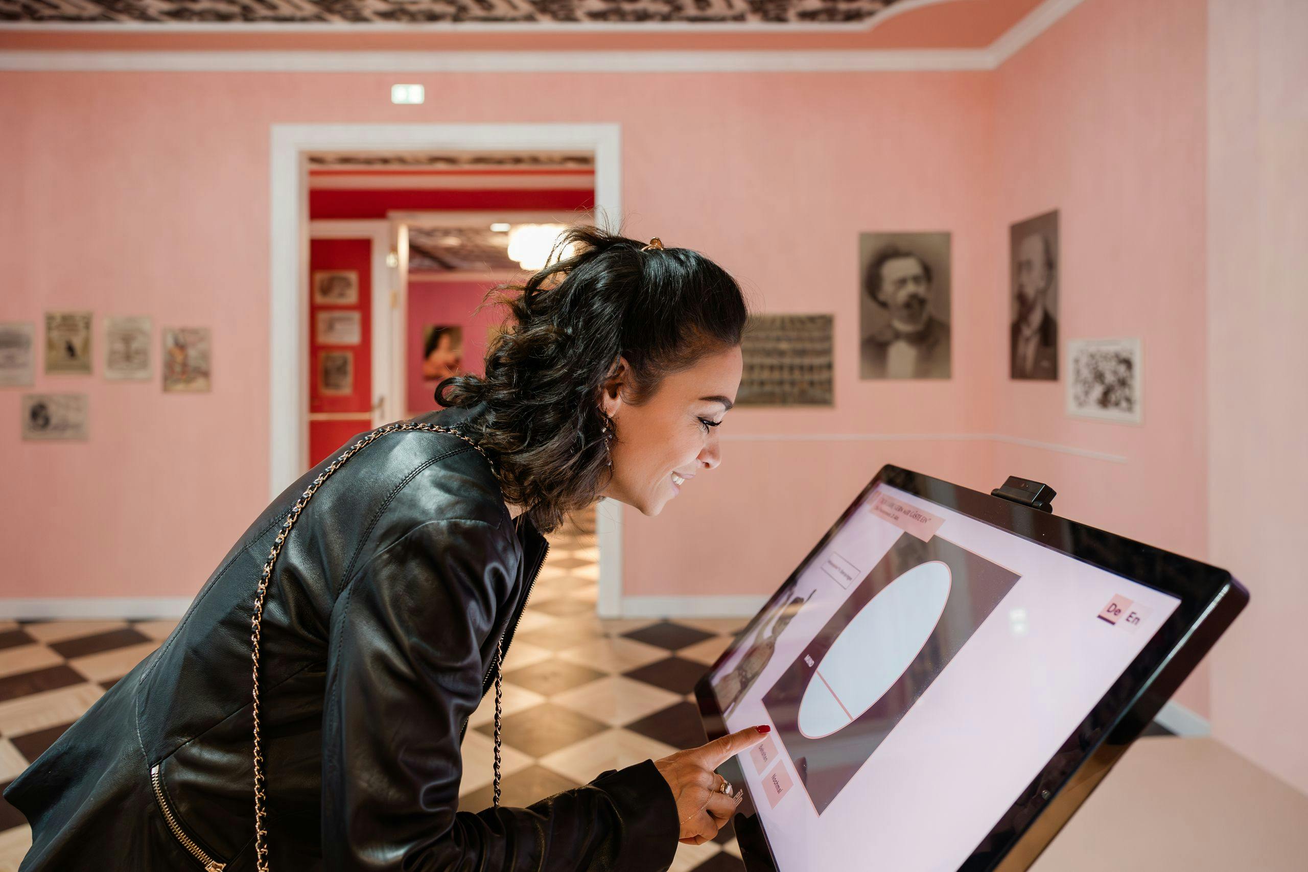 Vrouw in zwart leren jasje interageert met een touchscreen tentoonstelling in een roze kamer met ingelijste foto's aan de muren.