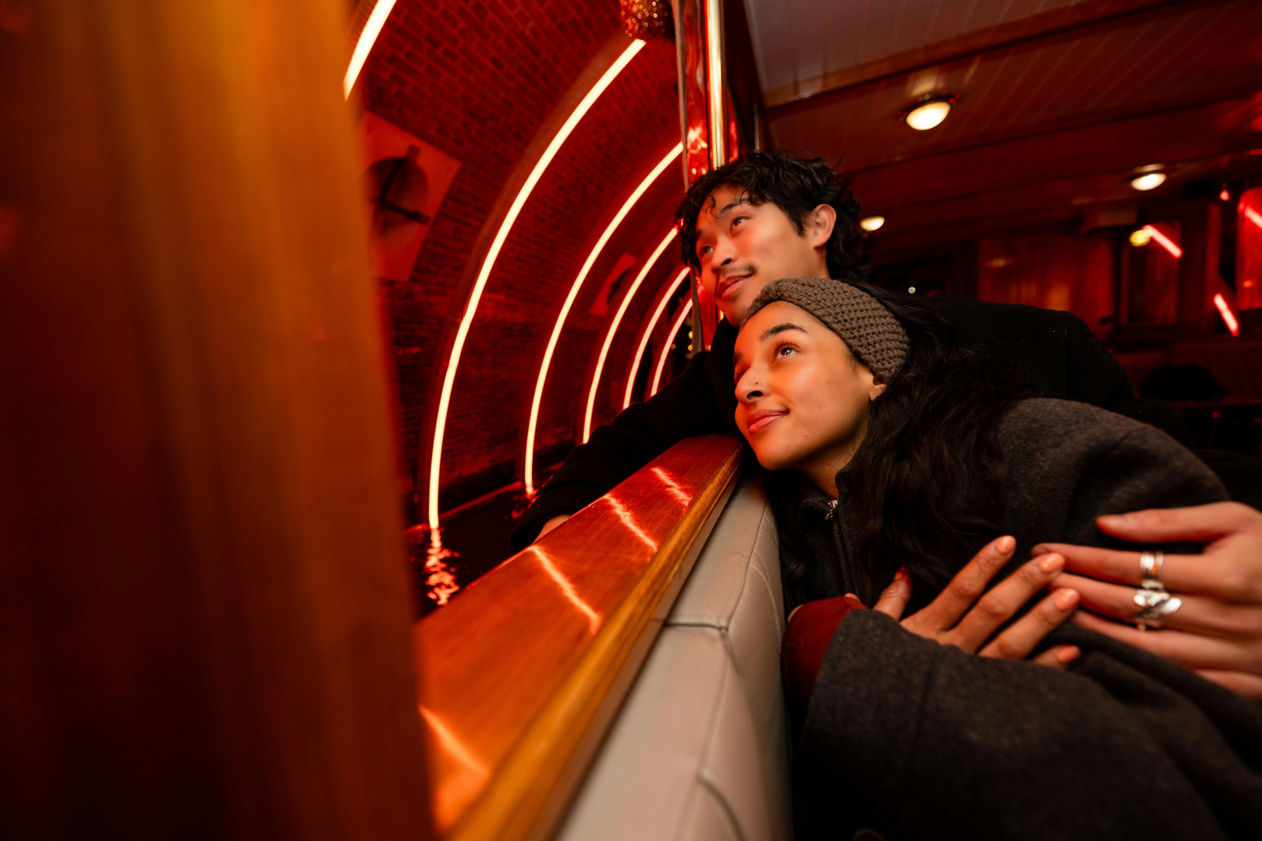 Zwei Personen lehnen an der Reling eines Bootes, lächeln und blicken hinaus auf rot beleuchtete Bögen über dem Wasser.