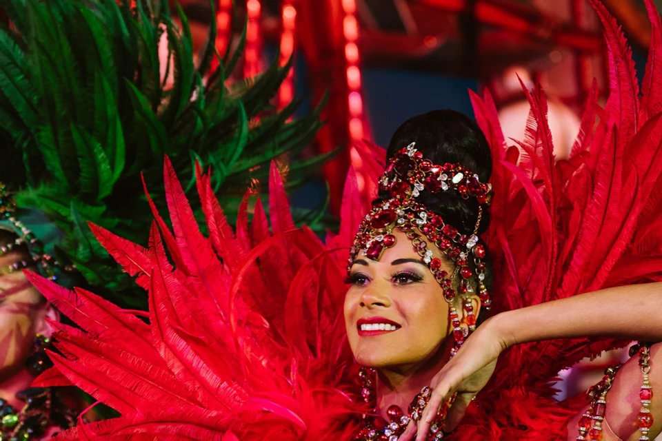 Performers in elaborate feathered costumes with red, green, and blue elements, dancing on a vibrant stage.