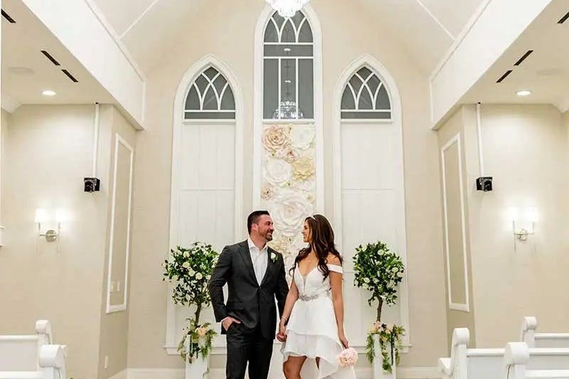 A couple, dressed in wedding attire, smiling and holding hands in a bright chapel with floral decorations and arched windows.