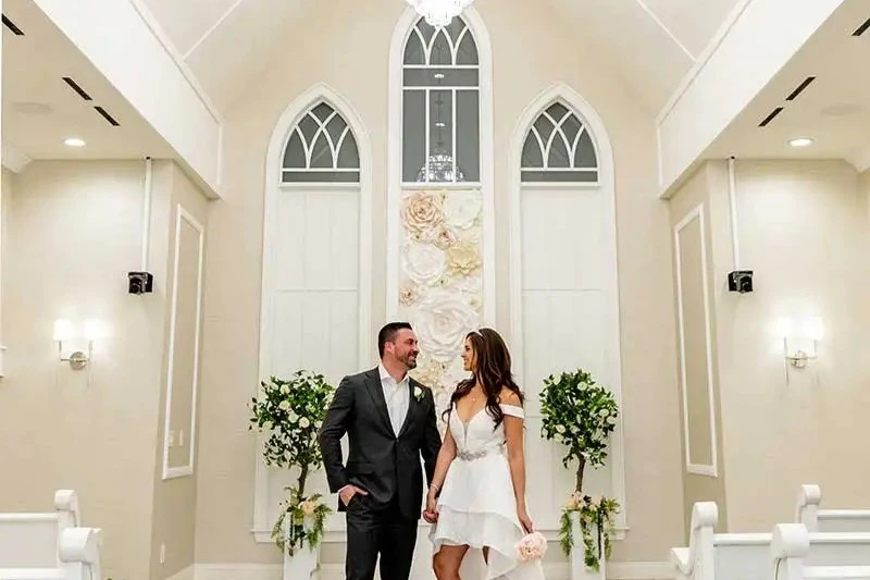A couple, dressed in wedding attire, smiling and holding hands in a bright chapel with floral decorations and arched windows.