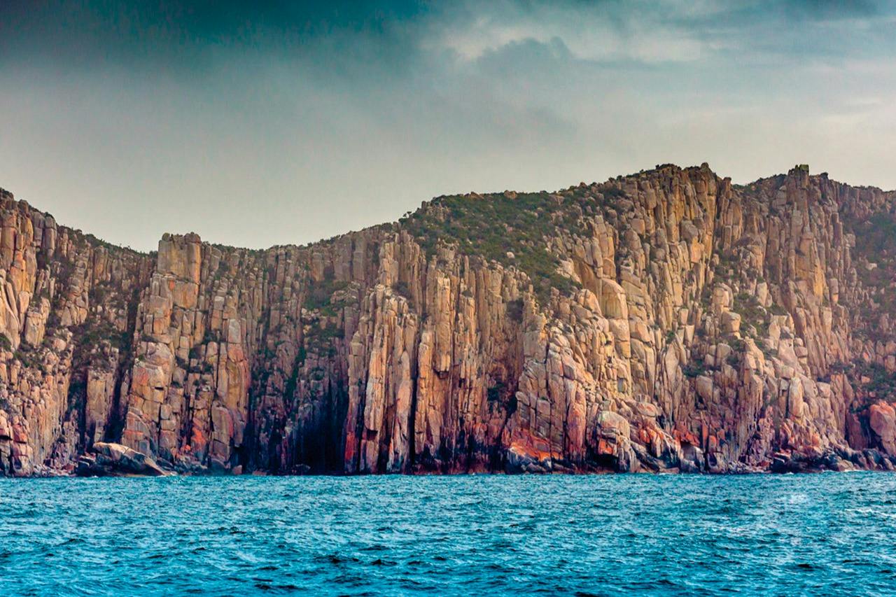 Rocky cliff face with sparse vegetation rising above blue ocean water under a cloudy sky.