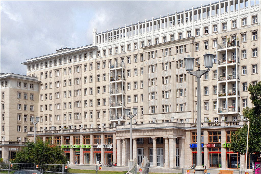 Multi-story beige building with balconies, large windows, and ground-floor shops under a cloudy sky. Lamp posts and trees in foreground.