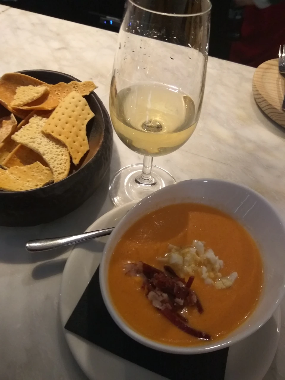 A bowl of orange soup with garnishes and a spoon next to a glass of white wine and a black bowl of crispy bread slices on a table.