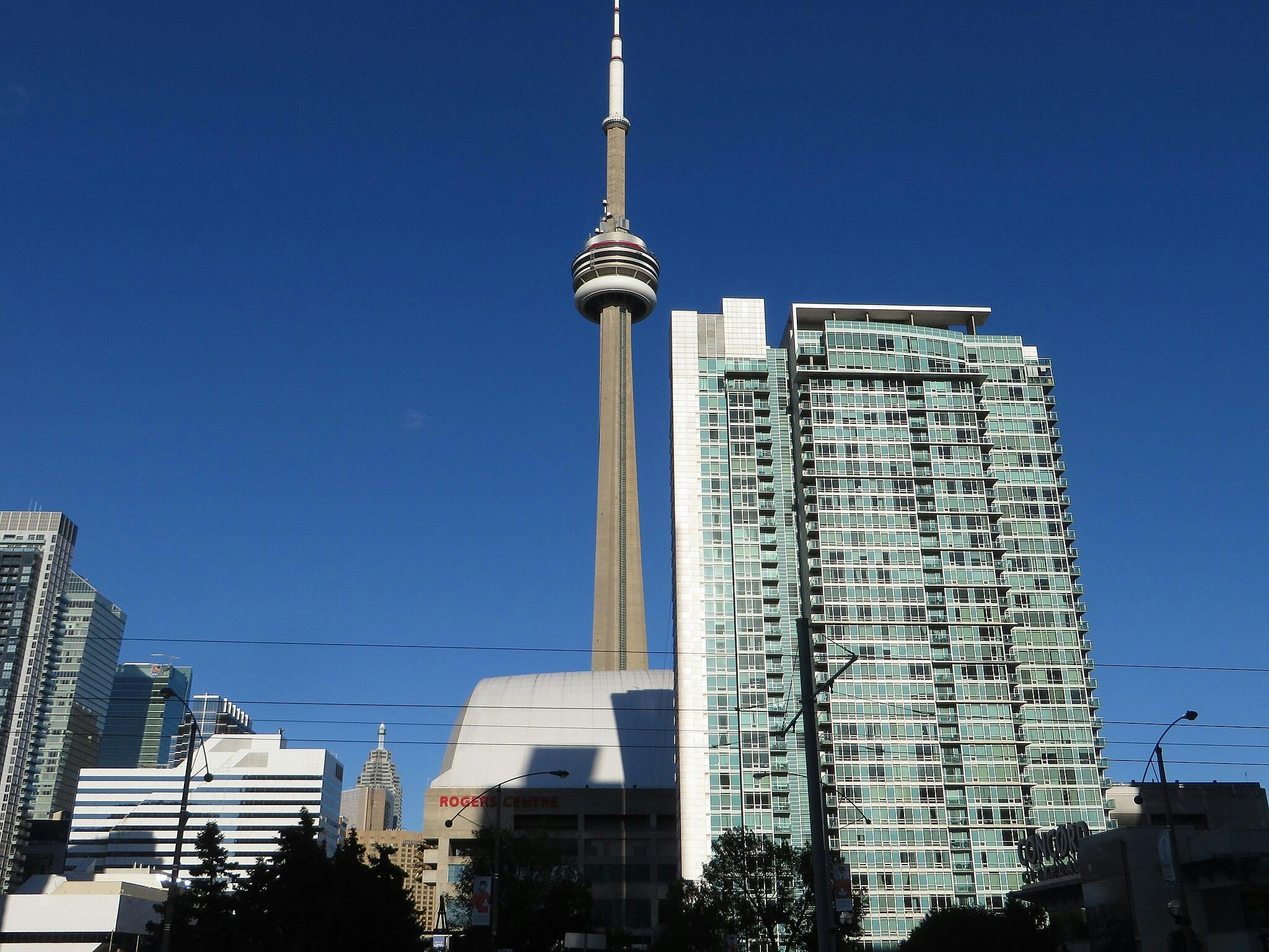 Hoher Turm mit Aussichtsplattform, umgeben von modernen Hochhäusern unter einem klaren blauen Himmel. Die Beschilderung "Rogers Centre" ist sichtbar.