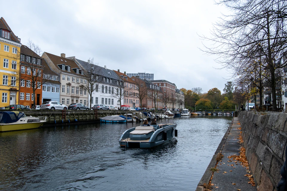 A canal with a small boat, lined with buildings and parked boats, under an overcast sky.