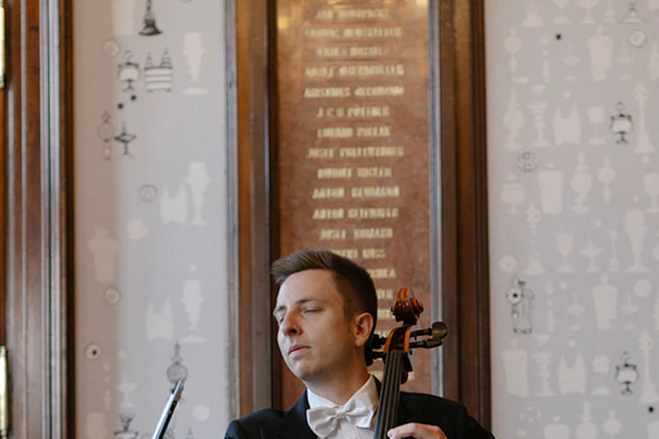 Four musicians in formal attire with string instruments walk through a grand hall with marble columns and arches.