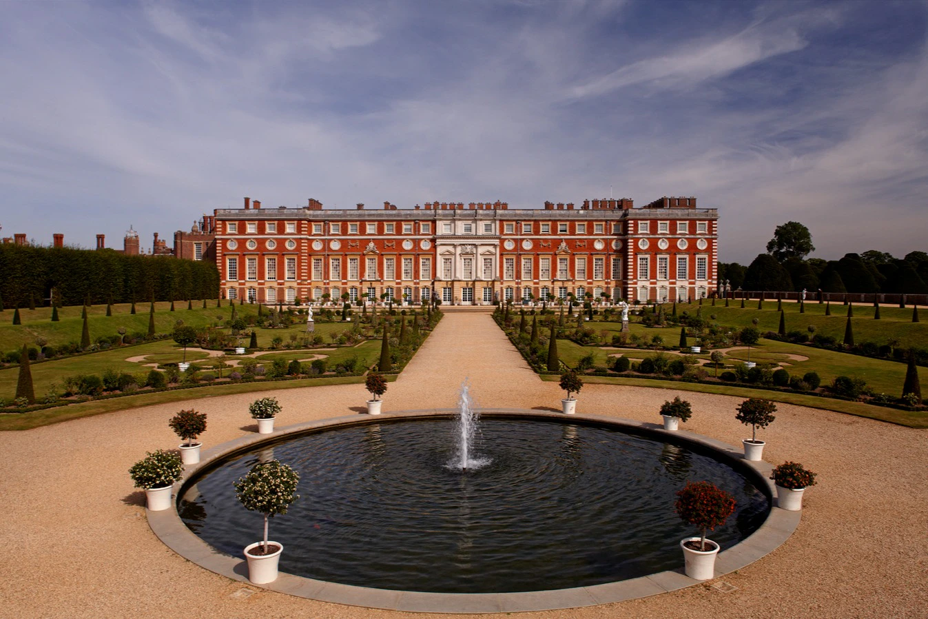 A large, ornate red-brick palace with a symmetrical garden and central fountain under a partly cloudy sky.