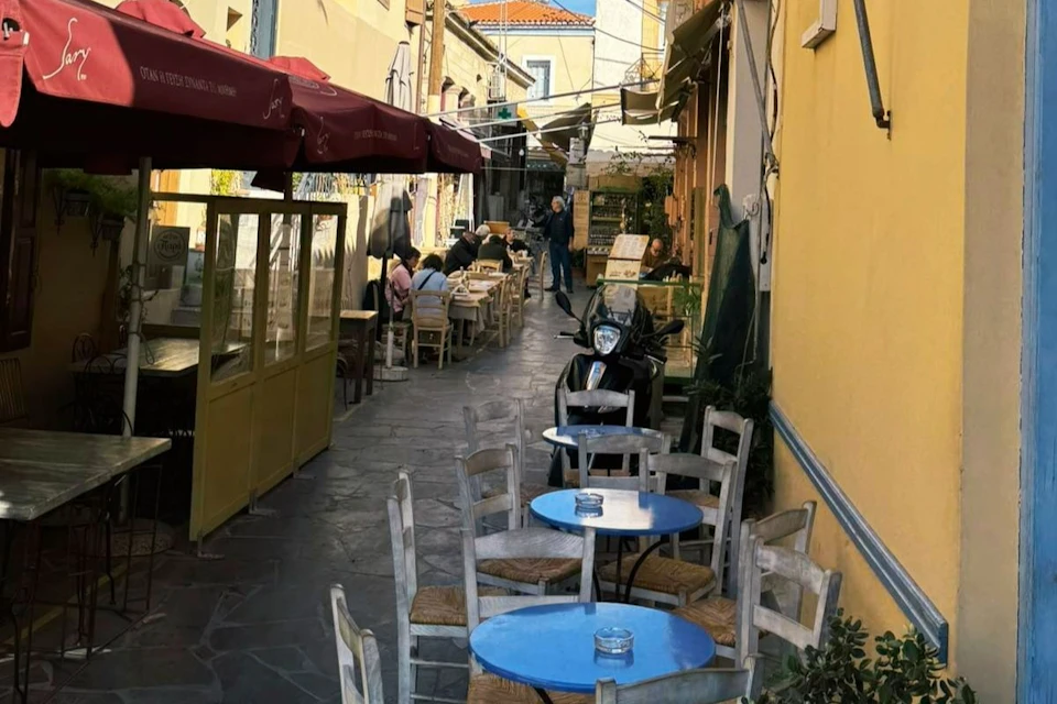 Outdoor café with blue tables and wooden chairs lining a narrow alleyway. People are seated in the background under umbrellas.