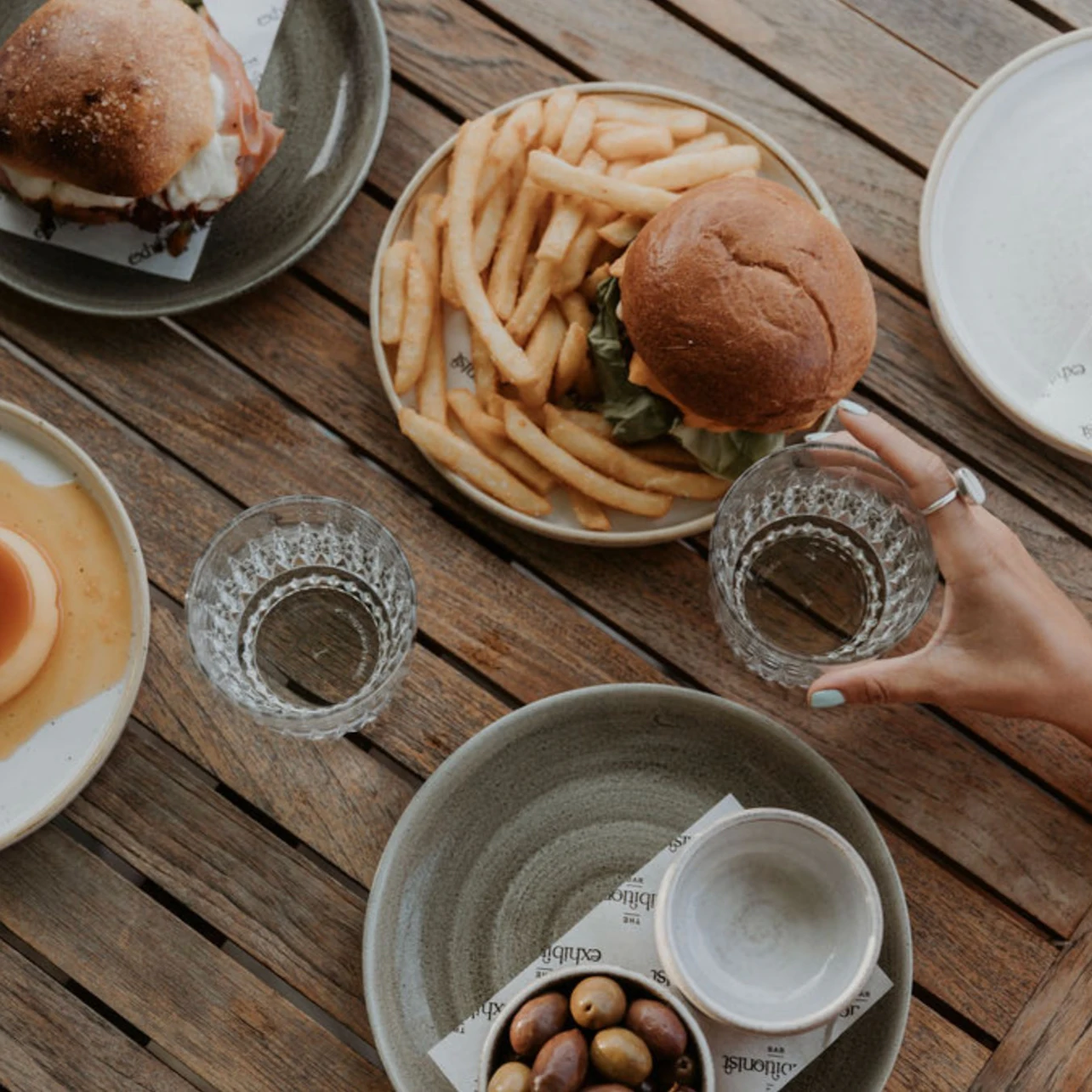 A table setting with burgers, fries, a dessert, olives, and glasses of water. A hand reaches for a glass.