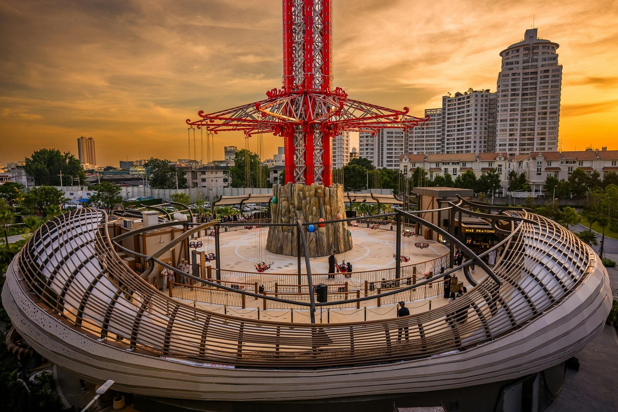 A tall amusement park ride with a circular base surrounded by buildings, under a vibrant orange evening sky.
