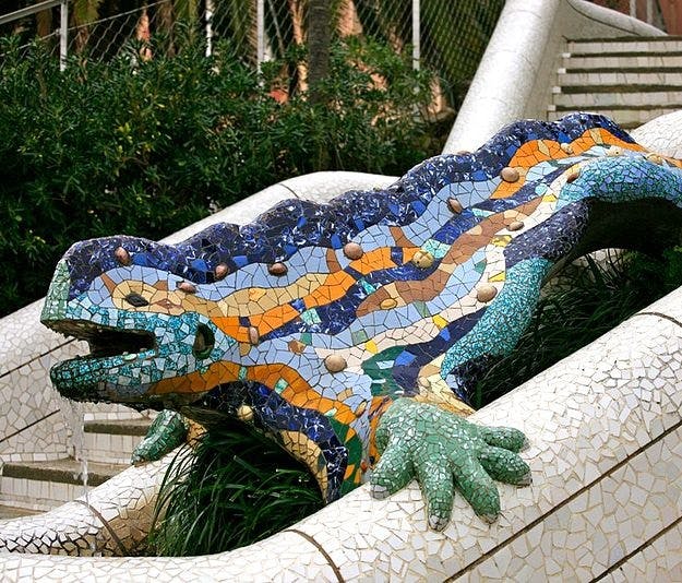 A colorful mosaic lizard sculpture situated on a white-tiled structure with greenery and stairs in the background.