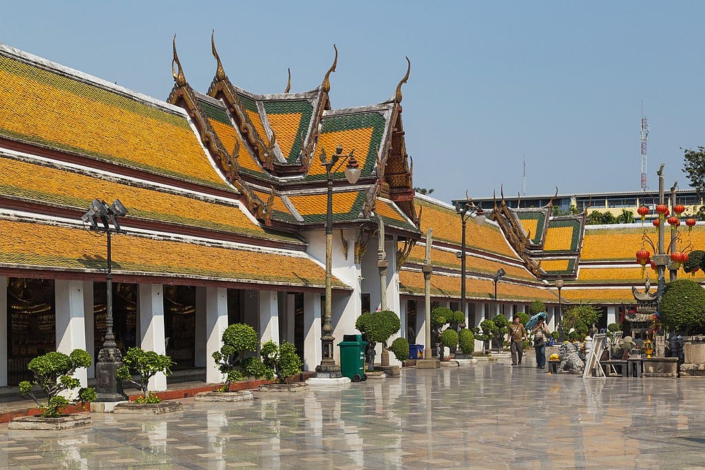 Persones passejant per un pati amb edificis adornats amb sostre daurat i plantes ben cuidades en un dia assolellat.