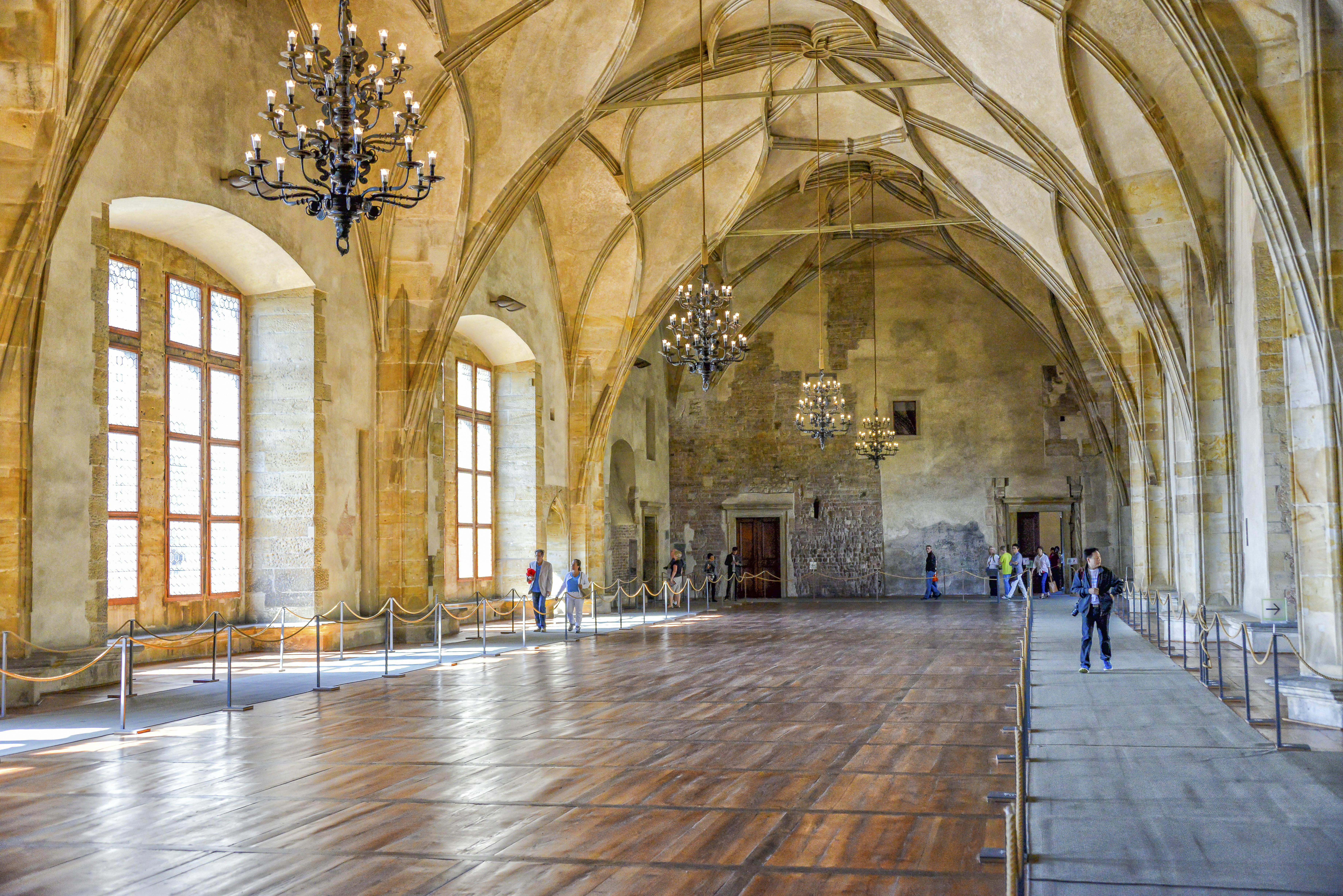 A grand hall with a vaulted ceiling, chandeliers, and large windows. Several people are walking or standing along the roped-off sides.