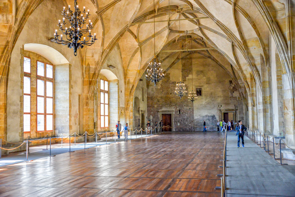 A grand hall with a vaulted ceiling, chandeliers, and large windows. Several people are walking or standing along the roped-off sides.