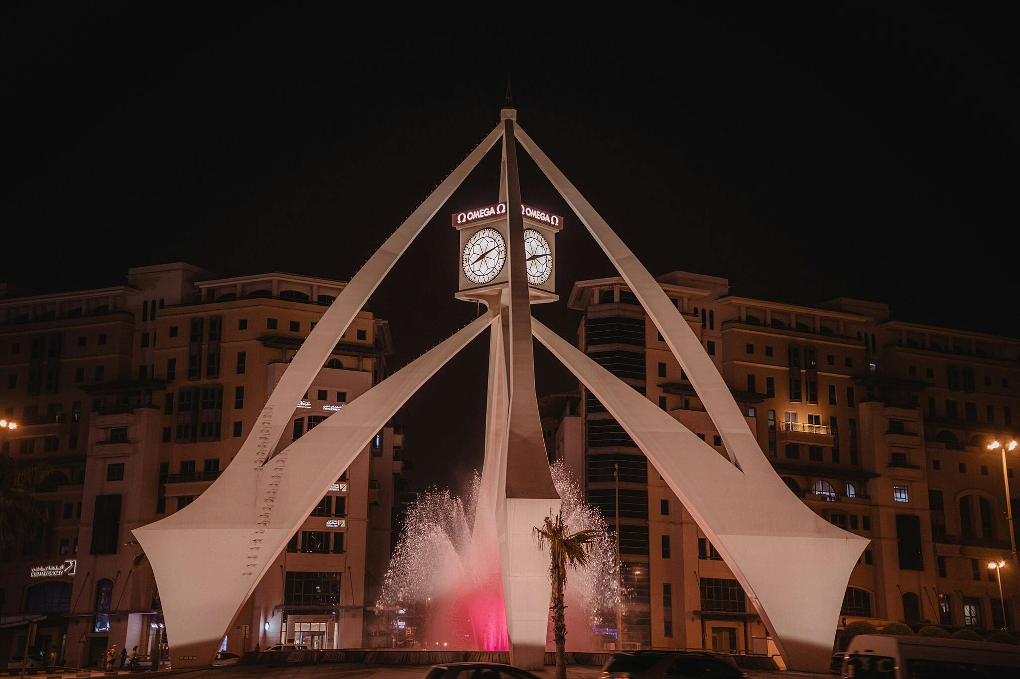 A clock tower with illuminated faces and "OMEGA" signs, surrounded by an arch structure and a fountain, located in front of buildings at night.