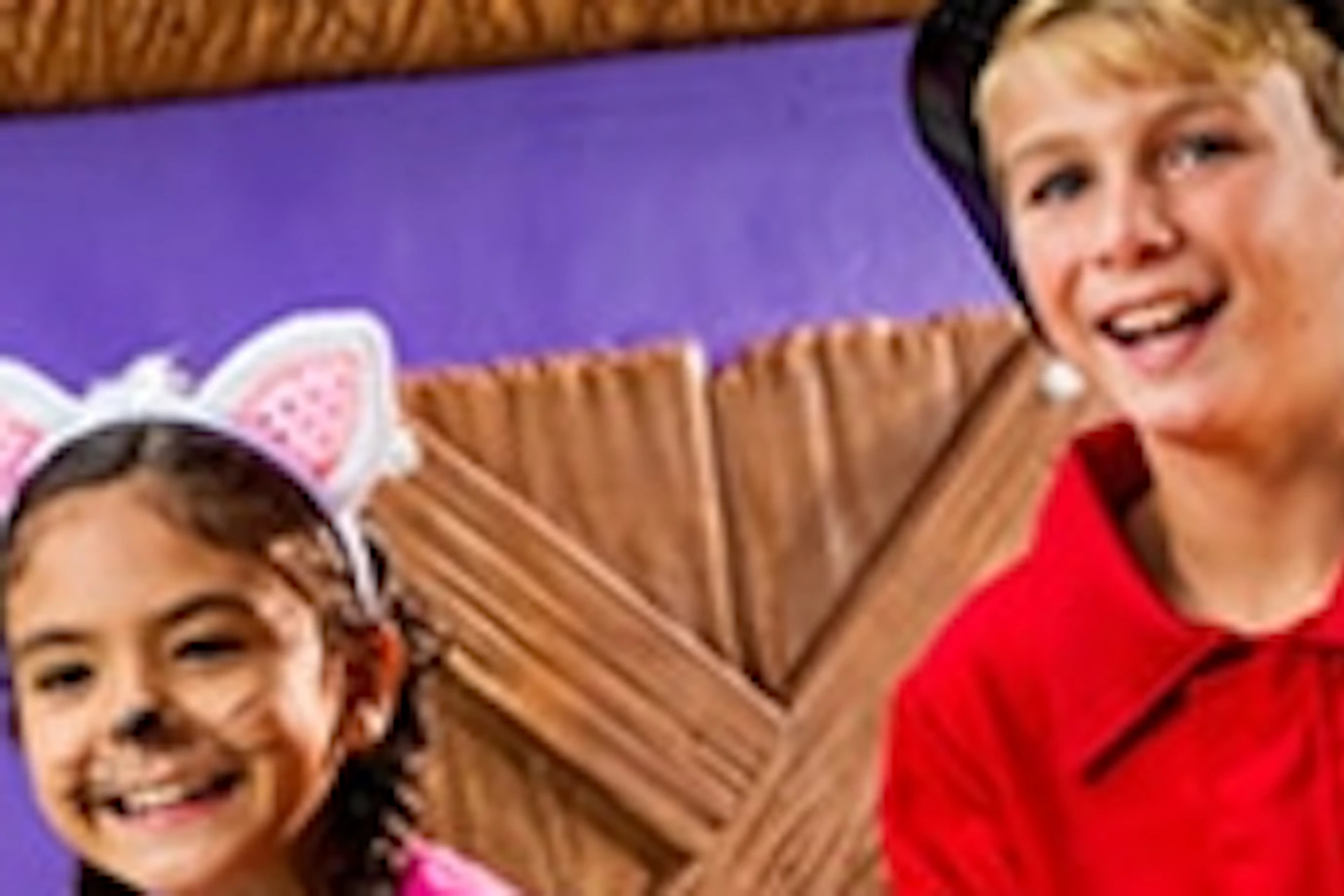 Two children in Halloween costumes, a pink cat and a firefighter, holding candy buckets in front of a playful, spooky house.