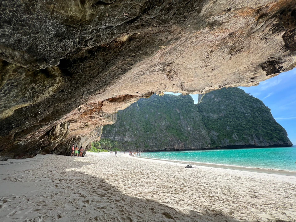 Sandy beach under a large rock overhang, with turquoise water and distant green cliffs. Several people are visible on the beach.