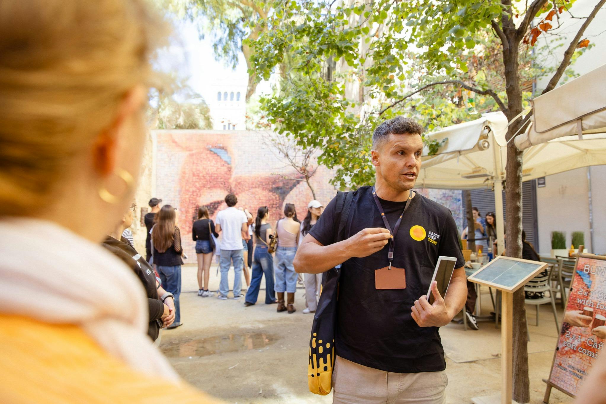 Un homme parlant dans un cadre extérieur avec des arbres et des gens en arrière-plan, tenant une tablette et faisant des gestes.