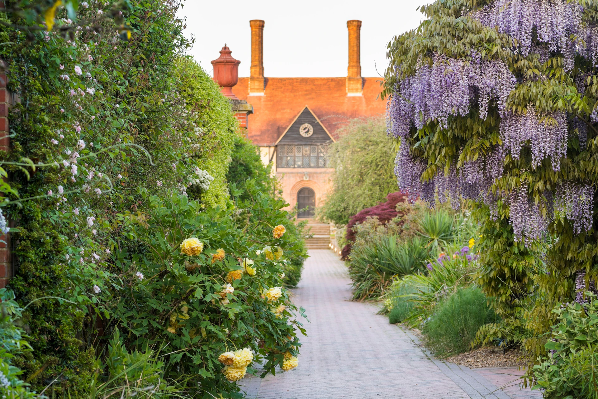 Pathway flanked by lush greenery and blooming flowers leads to a historic building with red chimneys and a triangular roof.