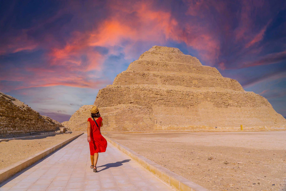 A person in a red dress and hat walks towards a step pyramid under a vivid sunset sky.