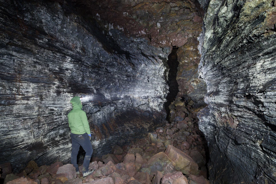 Person in a green jacket and dark pants stands in a rocky cave, illuminated by a headlamp.