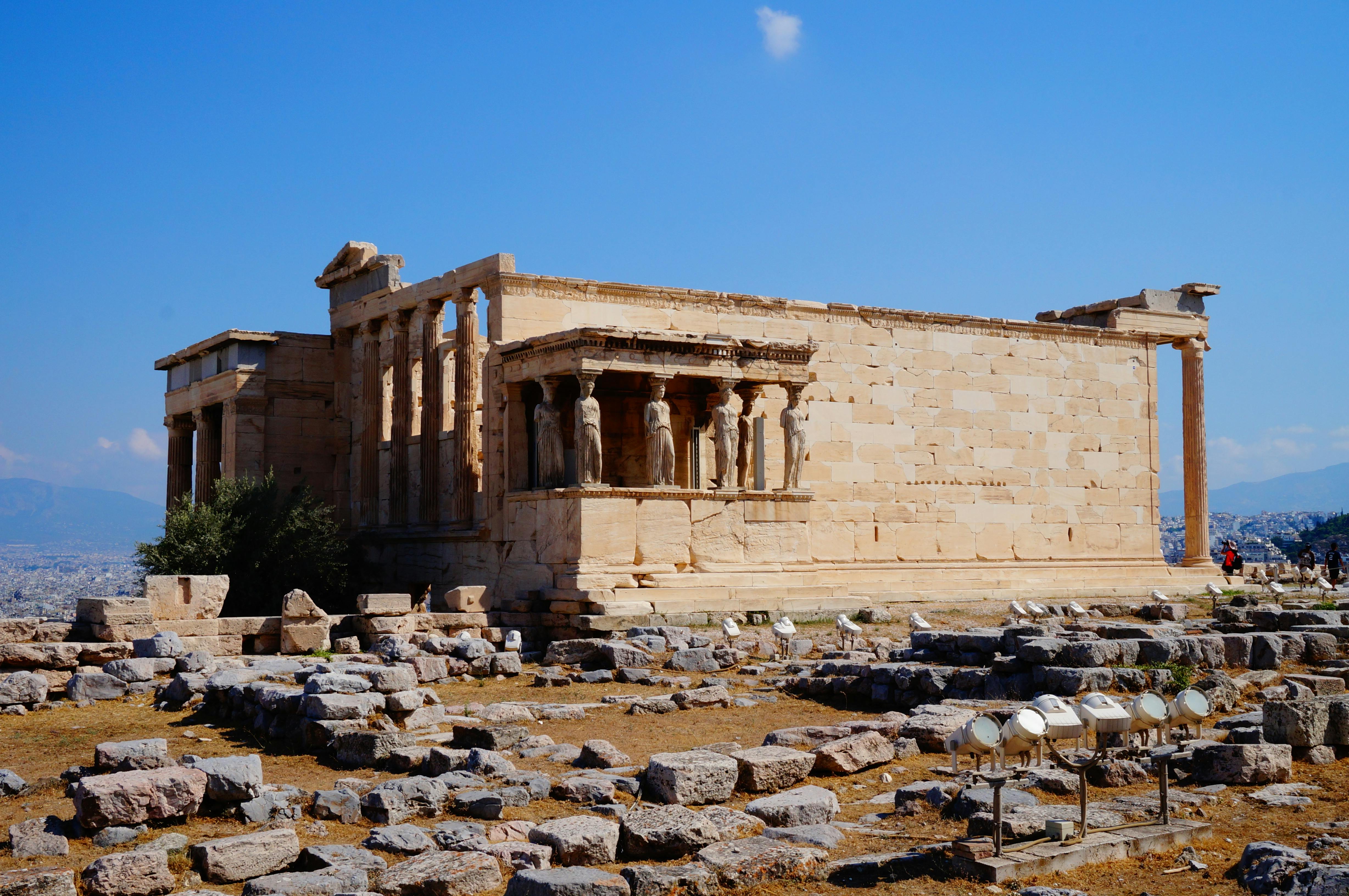 Ancient Greek temple with statues of women as pillars, surrounded by scattered rocks and ruins under a clear blue sky.