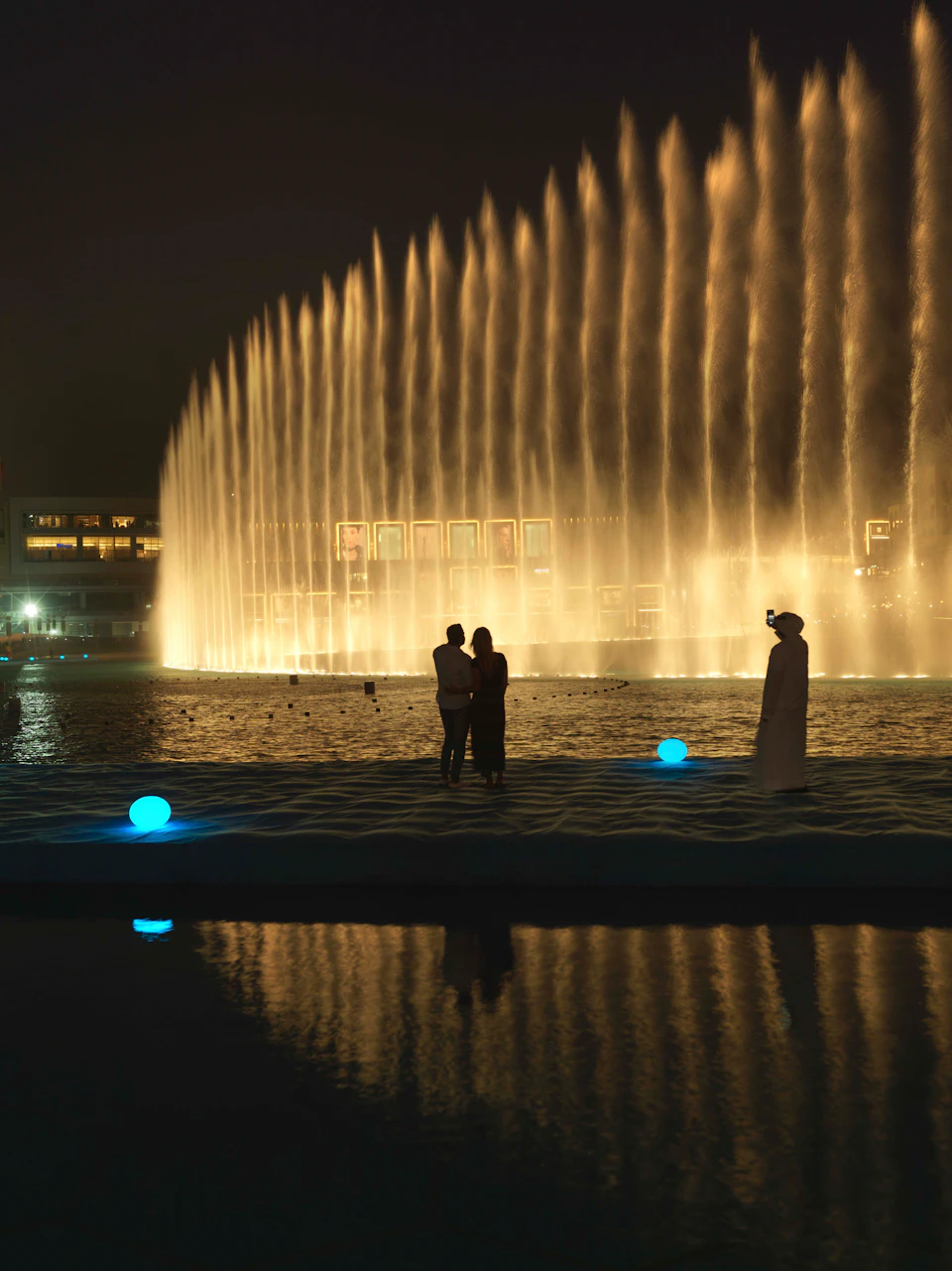 A couple is silhouetted against an illuminated fountain at night while a man in white robes films them.