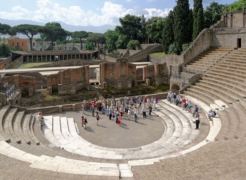 A group of tourists gathers in the ancient, partially restored amphitheater at Pompeii, surrounded by ruins and greenery.